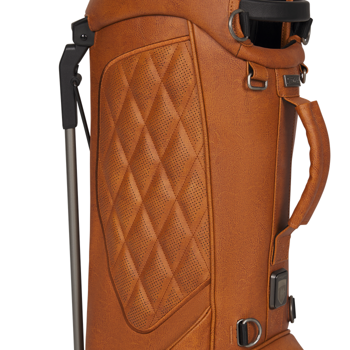 Close-up of a tan leather Titleist LINKSLEGEND Classic Members II Golf Stand Bag with quilted diamond stitching on the front panel, black hardware, a top carry handle, and part of a metal golf club shaft visible on the left.