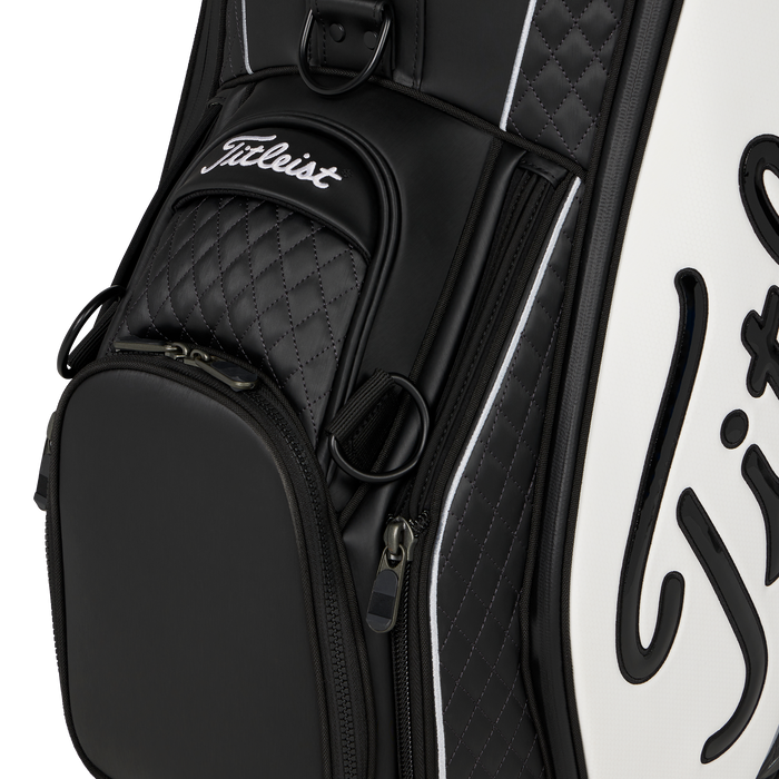 Close-up of a Titleist Tour Series Tour Golf Bag II in black and white, showing quilted black panels, a front zip pocket, and white script branding on a clean white background.