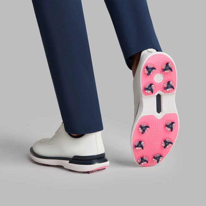 G/FORE Gallivan2R Split Toe G/LOCK Golf Shoes in white with navy trim and pink sole. View on a model lifting one shoe up showing pink sole 