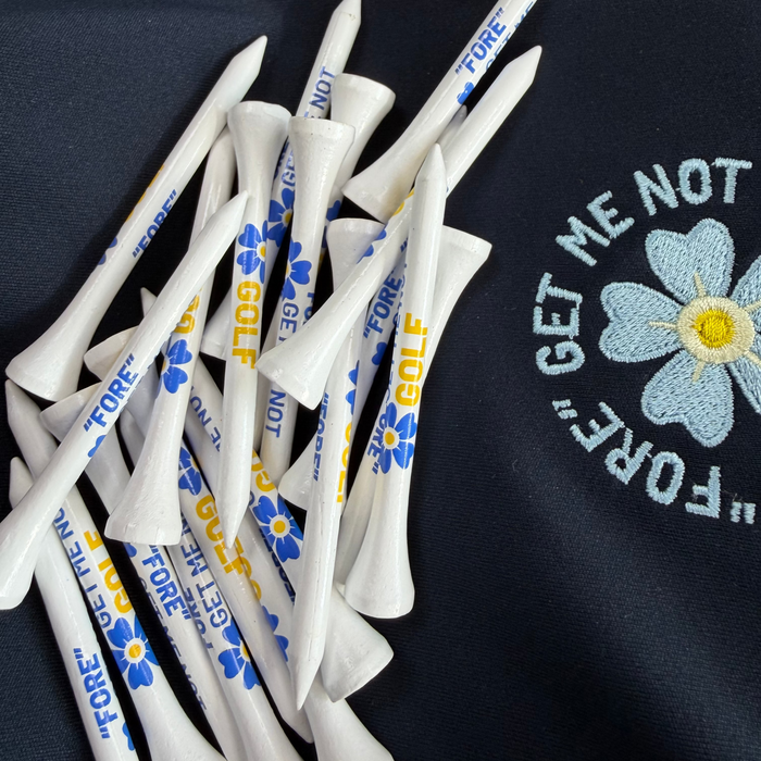 White bamboo Fore Get Me Not golf tees with blue flower and yellow text detail, scattered on a dark navy fabric background.