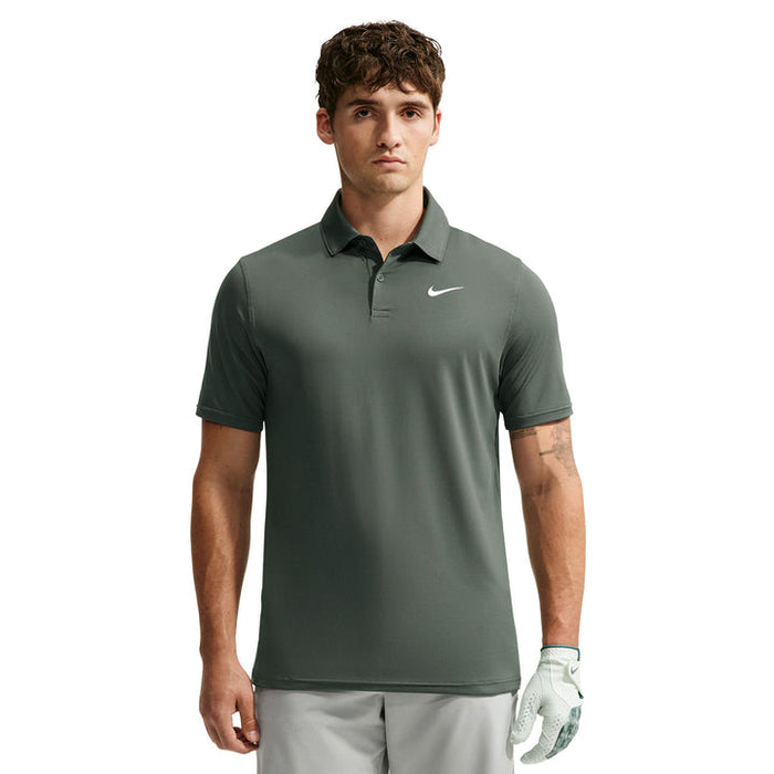 A man wearing a dark grey Nike polo shirt with a white swoosh logo on the chest, paired with light grey golf pants and a white golf glove, set against a plain white background.