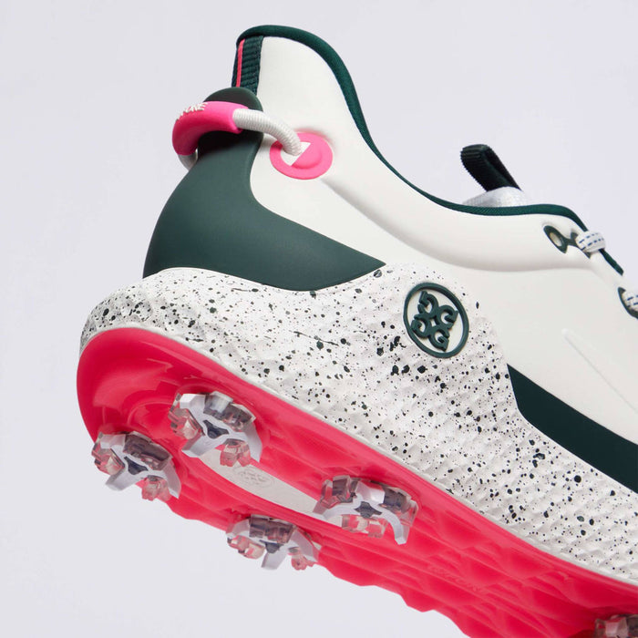 A close-up of the G/FORE MG4+ O2 G/LOCK Perforated Men's Golf Shoes showing the heel, midsole, and outsole. The shoe features a white upper with dark green overlays, a speckled white midsole, and a bright pink outsole with clear golf spikes.