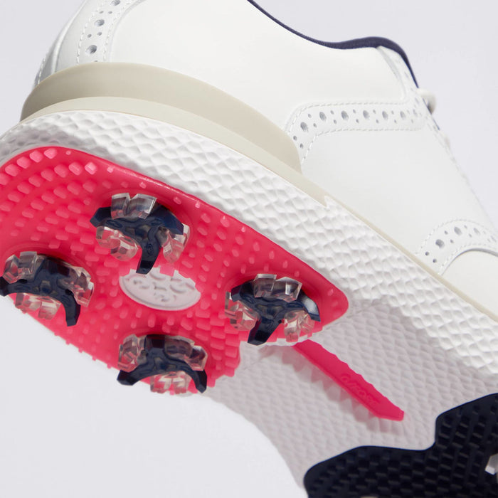 Close-up of a white G/FORE Gallivan2R Brogue Saddle Men's Golf Shoes showing the textured white upper, beige midsole, and bright pink sole with dark navy golf spikes.