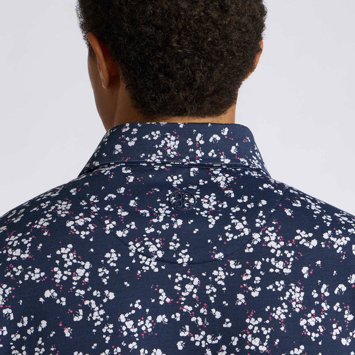 Back view of a man wearing a navy blue collared G/FORE Wild FlowerTech Jersey Men's Golf Polo Shirt with a dense white and pink floral print, shown against a plain light grey background.