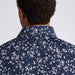 Back view of a man wearing a navy blue collared G/FORE Wild FlowerTech Jersey Men's Golf Polo Shirt with a dense white and pink floral print, shown against a plain light grey background.