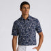 A man wearing a navy blue short-sleeve button-up G/FORE Wild FlowerTech Jersey Men's Golf Polo Shirt with a dense white floral print, paired with white pants, photographed against a light grey studio background.
