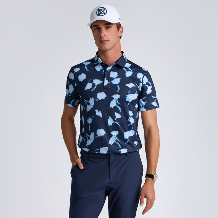 Man wearing a navy blue G/FORE Ghost Floral Tech Jersey Men's Golf Polo Shirt and white cap on a light grey background