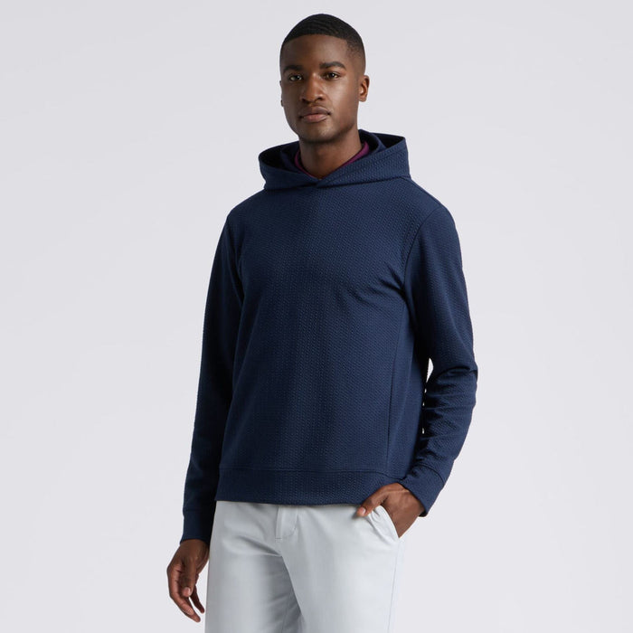 A man wearing a navy blue G/FORE Pebble Quilted Men's Golf Hoodie with a subtle waffle or honeycomb pattern, paired with light grey trousers, photographed against a solid light grey background.