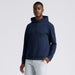 A man wearing a navy blue G/FORE Pebble Quilted Men's Golf Hoodie with a subtle waffle or honeycomb pattern, paired with light grey trousers, photographed against a solid light grey background.