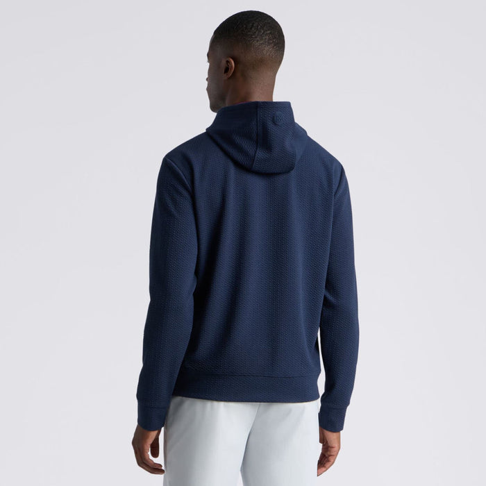A man viewed from the back wearing a textured navy blue G/FORE Pebble Quilted Men's Golf Hoodie and white pants, set against a plain light grey studio background.