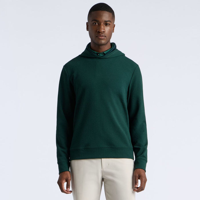 A man wearing a dark green knit G/FORE Pebble Quilted Men's Golf Hoodie with a high folded collar, paired with light beige trousers, standing against a light grey studio background.