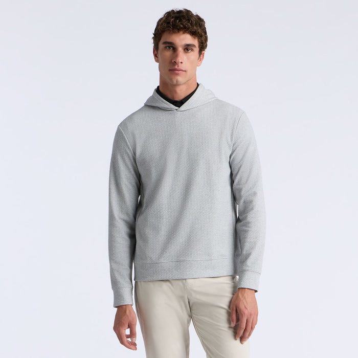 A man wearing a light grey textured hooded G/FORE Pebble Quilted Men's Golf Hoodie over a black shirt and beige pants, standing against a plain white studio background.