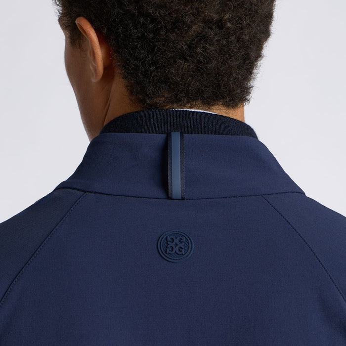 Close-up back view of a person wearing a navy blue G/FORE Storm Fleece Hybrid Men's Golf Jacket, showing the collar, shoulder seams, and a circular tonal logo patch on the upper back against a light grey background.